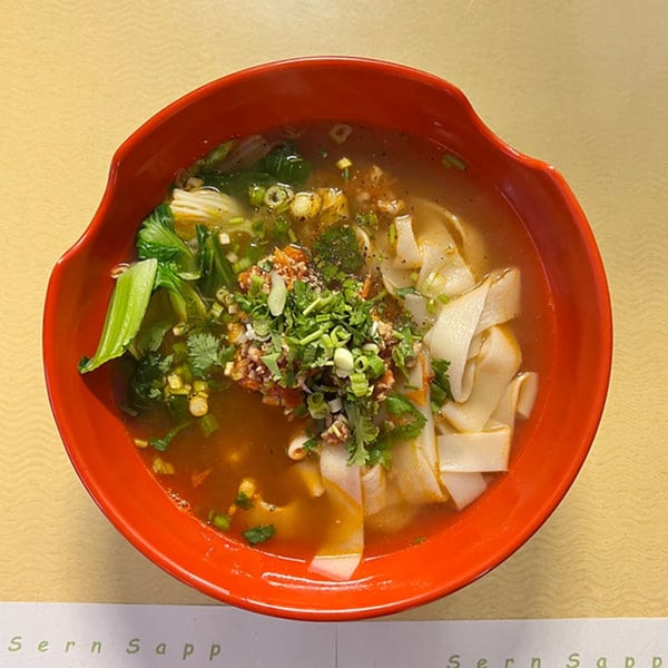 A vibrant red bowl of Sern Sapp Restaurant's Laos noodle soup with bok choy, herbs, and spices.